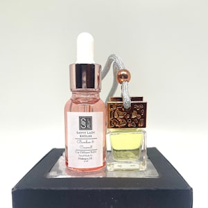 May include: A small glass bottle with a rose-colored liquid and a gold cap. The bottle is labeled "Savvy Lady Kndles Bamboo & Coconut Car Diffuser Refill Hand Made in Makan M". The bottle is next to a small glass car air freshener with a gold top and a gray string.