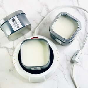 May include: Three square candle tins, two are silver and one is on a white electric warmer. The silver tins have a label that says "Simply Lit Candles" and "Vanilla & Coconut".