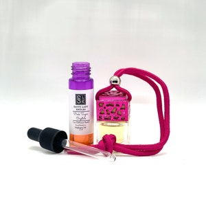 May include: A pink and purple car air freshener diffuser with a pink string. The bottle is labeled "Savvy Lady Candles Pink Sugar Crystals Car Diffuser Refill Hand Made in Michigan, MI 15 ml".