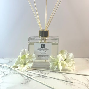 May include: A clear glass bottle with a gold lid and a label that reads "Savvy Lady Kndles Pink Sugar Crystals Reed Diffuser Handmade Muskegon, Mi 495/113-48". The bottle is filled with a clear liquid and has wooden sticks sticking out of the top. The bottle is sitting on a white marble surface with white flowers.