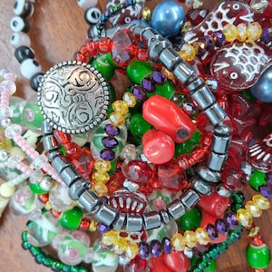 May include: A collection of colorful beaded necklaces and bracelets. The jewelry features a variety of beads in red, green, purple, yellow, and black, with silver-toned metal accents. Some beads have unique shapes, including fish and coral.