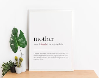 May include: A white framed print with the word "mother" in black text. The definition of the word is in smaller black text, and the name "Angela" is in red text. The print is sitting on a wooden surface with a green plant and a cactus in a white pot.