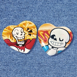 May include: Two heart-shaped pins featuring characters from the video game Undertale. One pin shows Papyrus with spaghetti, the other shows Sans with a hot dog. The pins are set against a blue denim background.