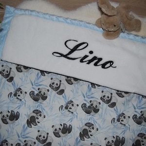 Peut inclure: Une couverture de bébé blanche avec une bordure bleue et un imprimé panda. La couverture a un rabat blanc avec le nom "Lino" brodé en noir.