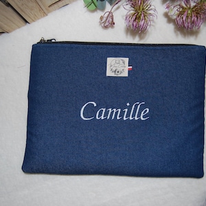Peut inclure: Une pochette rectangulaire en tissu bleu foncé dotée d'une fermeture éclair noire sur le bord supérieur. Le prénom Camille est brodé en blanc en lettres cursives au centre. Une petite étiquette blanche est cousue au-dessus du nom.
