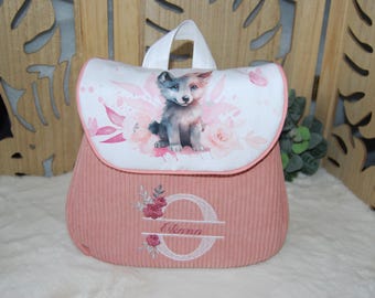 Könnte beinhalten: Ein altrosa Rucksack aus Kord mit einer weißen Klappe, die eine Aquarellillustration eines Wolfswelpen umgeben von rosa Blumen zeigt. Auf der Vorderseite befindet sich ein gestickter Buchstabe O mit dem Namen Oliana und Blumenmotiven.