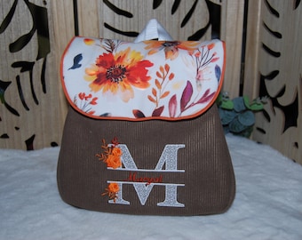 Könnte beinhalten: Ein brauner Rucksack aus Kord mit einer weißen Klappe, die mit einem leuchtend orangefarbenen und roten Blumenmuster bedruckt ist. Vorne befindet sich ein großer, gestickter Buchstabe M in gemusterter grauer Schrift, verziert mit kleinen orangefarbenen Blüten und dem Namen Margot in Rot.