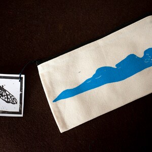 Könnte beinhalten: Cremefarbene Canvas-Reißverschlusstasche mit blauem, abstraktem Design. Ein kleines Etikett mit einem schwarz-weißen Schmetterlingsdruck ist angebracht. Die Tasche hat einen schwarzen Reißverschluss und einen schwarzen Zipper. Der Hintergrund ist dunkelbraun.