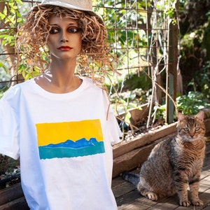 Può includere: Una maglietta bianca con un design a paesaggio giallo, blu e verde. La maglietta è indossata da un manichino con un cappello di paglia. Un gatto tigrato è seduto su una superficie di legno sullo sfondo.