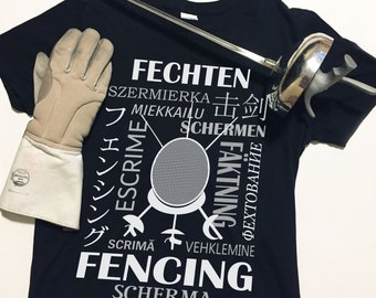 May include: Black t-shirt with a white graphic design featuring a fencing mask and the words "FECHTEN", "SZERMIERKA", "MIEKKAILU", "ESCRIME", "FÄKTNING", "SCRIMÃ", "VEHKLEMINNE", "FENCING", and "SCHERMA" in different languages.