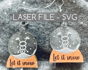 May include: A pair of laser cut wooden earrings with a clear acrylic snow globe charm. The snow globe features a white snowman with arms outstretched and snowflakes. The wooden base of the earrings is engraved with the text "Let it snow".