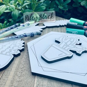 Puede incluir: Un letrero hexagonal de madera blanca con un contorno negro de un cactus en una maceta. El letrero est&aacute; sobre una superficie de madera con marcadores verdes y un letrero que dice "Riley Black Made with Love & A Little Sass".