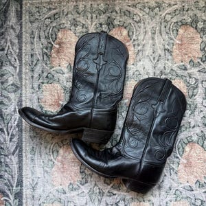 Könnte beinhalten: Ein Paar schwarze Cowboystiefel aus Leder mit aufwendigen, farblich abgestimmten Stickmustern auf dem Schaft, spitzer Zehenpartie, Blockabsätzen und Zuglaschen am oberen Rand, die eine klassische Silhouette mit detaillierten Stickereien zeigen.