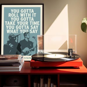 Puede incluir: Un póster enmarcado con el texto "YOU GOTTA ROLL WITH IT" y una ilustración de banda está sobre un mueble rojo. Un tocadiscos con un disco de vinilo negro está sobre el mueble. Libros apilados frente al póster.