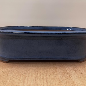 May include: A rectangular bonsai pot with a dark blue glaze. The ceramic container has a rounded rim and sits on four small feet. The interior is a contrasting brown color. This container is suitable for bonsai trees.