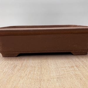 May include: A rectangular, brown bonsai pot with a slightly textured surface. The pot has a low profile with a flat bottom and four small feet. The sides are straight, and the rim is slightly rounded. Ideal for bonsai trees.