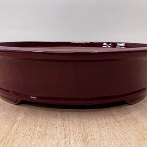 May include: A burgundy-colored, oval-shaped bonsai pot. The ceramic pot has a glossy finish and a slightly raised rim. The pot sits on three small feet. The interior of the pot is a lighter brown color.