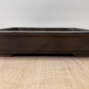 May include: A rectangular, dark brown bonsai pot with a slightly flared rim and four small feet. The ceramic pot has a smooth, matte finish and is designed for planting small trees or shrubs. The pot is approximately 12 inches long.