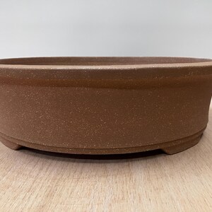 May include: A brown, oval-shaped bonsai pot with a textured surface. The pot has a wide, shallow design with a slightly flared rim and a flat base. The pot is made of ceramic or a similar material.