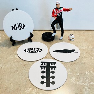 May include: A collection of white coasters with black graphics and text, including the letters "NHRA" and a drag racing car silhouette. A miniature race car driver figure stands near a tire and a helmet. The coasters are on a light-colored surface.