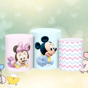 May include: Three cylindrical decorations. One is pink with Minnie Mouse, another is light blue with Mickey Mouse, and the third has a pink and blue chevron pattern. The decorations are for a baby shower or birthday.