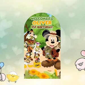 以下が含まれることがあります： ミッキーマウスがサファリ風の衣装を着て、他のディズニーキャラクターに囲まれた、カラフルな誕生日をテーマにしたグラフィック。 「WELCOME TO OLIVER 1ST BIRTHAY」の文字がキャラクターの上に、緑色の背景に表示されています。