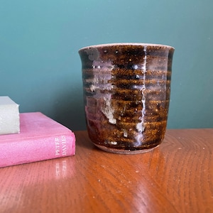 May include: A brown ceramic mug with a speckled glaze and a slightly flared rim. The mug is sitting on a wooden surface.