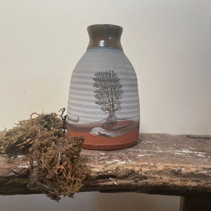 May include: A small, white ceramic vase with a brown rim and a tree design. The vase has a textured surface and a brown and white stripe at the bottom.