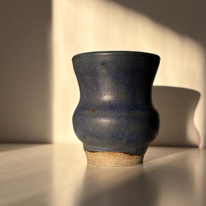 May include: A dark blue ceramic vase with a distinctive, curved form. The vase has a textured finish and a light beige base. The interior is not visible. The vase is likely used for decorative purposes.