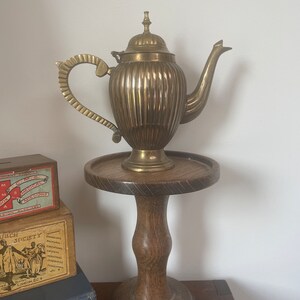 May include: A gold-coloured brass teapot with a fluted body and a decorative handle sits on a wooden pedestal. The teapot has a domed lid with a finial.