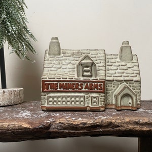 May include: A ceramic model of a pub called "The Miners Arms" with a stone facade and a red sign. The model is on a wooden surface.