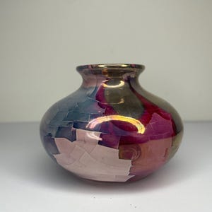 May include: A small, round ceramic vase with a crackled glaze in shades of pink, blue, and gold. The vase has a wide mouth and a narrow neck.