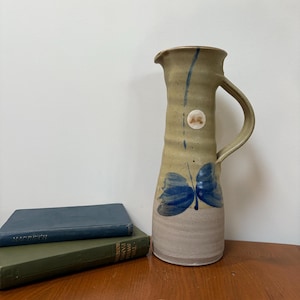 May include: A green and brown ceramic jug with a blue butterfly design. The jug has a handle and a spout. It is sitting on a wooden table with two books.