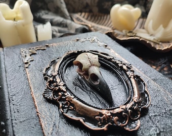 May include: A decorative, antique-style book with a framed bird skull centerpiece. The frame has an ornate, bronze-colored finish, set against a textured, dark gray surface. Two lit candles are in the background, creating a gothic aesthetic.