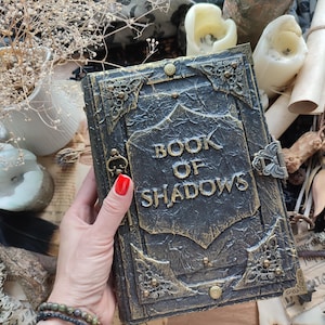 Może przedstawiać: Czarna i złota dekoracyjna książka z wytłoczonymi słowami "BOOK OF SHADOWS" na okładce. Książka ma ozdobne metalowe narożniki i zapięcie. Tło obejmuje świece, suszone kwiaty i pióro.