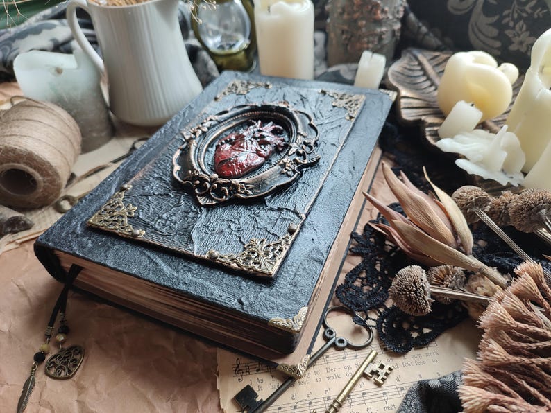 Pu&ograve; includere: Un diario rilegato in pelle nera con un design decorativo a forma di cuore rosso e dorato sulla copertina. Il diario &egrave; chiuso e ha un aspetto usurato e vintage.