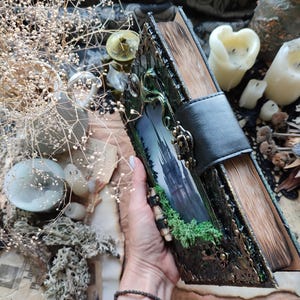 May include: A decorative, handcrafted book with a black leather strap closure. The cover features a landscape image of a castle and green foliage, with ornate metal accents. The aged pages and overall aesthetic suggest a fantasy or gothic theme.