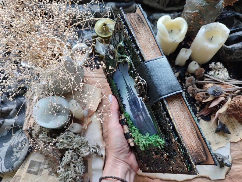 May include: A decorative, handcrafted book with a black leather strap closure. The cover features a landscape image of a castle and green foliage, with ornate metal accents. The aged pages and overall aesthetic suggest a fantasy or gothic theme.