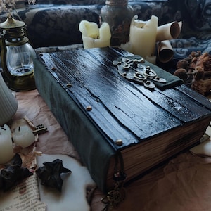Puede incluir: Un diario artesanal con una cubierta de madera oscura y un cierre decorativo de metal. El libro tiene una lomo y esquinas de ante verde, con tachuelas de latón. Un dije de llave decorativo cuelga de la columna vertebral. Las páginas están envejecidas.