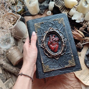 Pu&ograve; includere: Un diario rilegato in pelle nera con una cornice dorata ornata e un design a forma di cuore anatomico rosso. Il diario &egrave; chiuso e ha un aspetto usurato.