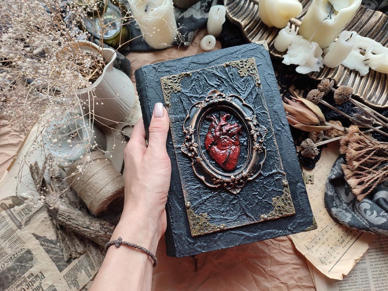 Pu&ograve; includere: Un diario rilegato in pelle nera con una cornice dorata ornata e un design a forma di cuore anatomico rosso. Il diario &egrave; chiuso e ha un aspetto usurato.