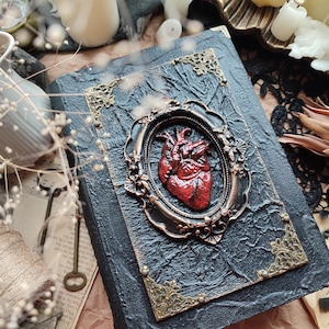 Pu&ograve; includere: Un diario rilegato in pelle nera con una cornice dorata ornata che circonda un cuore anatomico rosso. Il diario ha una superficie testurizzata e dettagli dorati.