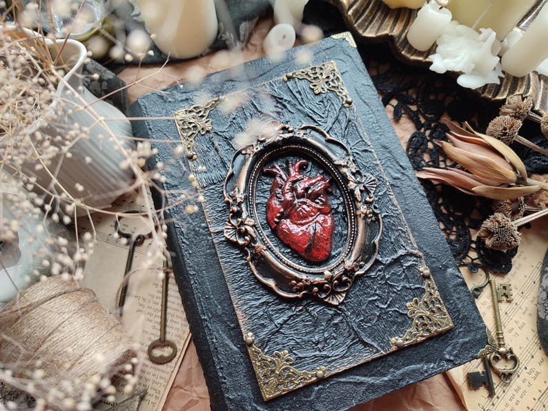 Pu&ograve; includere: Un diario rilegato in pelle nera con una cornice dorata ornata che circonda un cuore anatomico rosso. Il diario ha una superficie testurizzata e dettagli dorati.