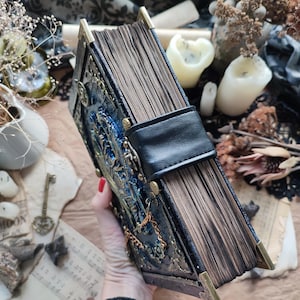 K&ouml;nnte beinhalten: Ein detailliertes Buch im antiken Stil mit einem dunkelblauen und goldenen Einband, der aufwendige gepr&auml;gte Designs und Metallecken aufweist. Das Buch hat einen schwarzen Lederriemenverschluss und gealterte, braun get&ouml;nte Seiten. Die &Auml;sthetik erinnert an ein Vintage-Grimoire oder -Journal.