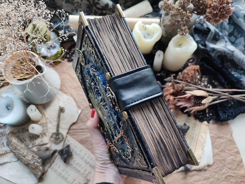 K&ouml;nnte beinhalten: Ein detailliertes Buch im antiken Stil mit einem dunkelblauen und goldenen Einband, der aufwendige gepr&auml;gte Designs und Metallecken aufweist. Das Buch hat einen schwarzen Lederriemenverschluss und gealterte, braun get&ouml;nte Seiten. Die &Auml;sthetik erinnert an ein Vintage-Grimoire oder -Journal.