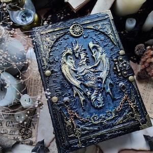 K&ouml;nnte beinhalten: Ein detailliertes, blau-goldenes Fantasy-Journal mit einem Drachendesign. Das Cover zeigt ein erhabenes Relief eines Drachen, einer Sonne und eines Mondes. Dekorative Ketten und Verschl&uuml;sse tragen zur gotischen &Auml;sthetik bei. Das Journal ist von Schl&uuml;sseln und Kerzen umgeben.