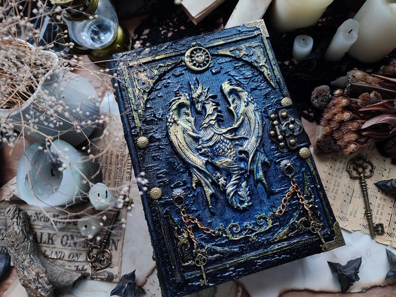 K&ouml;nnte beinhalten: Ein detailliertes, blau-goldenes Fantasy-Journal mit einem Drachendesign. Das Cover zeigt ein erhabenes Relief eines Drachen, einer Sonne und eines Mondes. Dekorative Ketten und Verschl&uuml;sse tragen zur gotischen &Auml;sthetik bei. Das Journal ist von Schl&uuml;sseln und Kerzen umgeben.