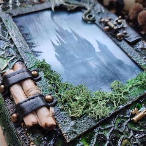 May include: A handcrafted, fantasy-themed picture frame with a castle scene. The frame is textured with green and brown tones, adorned with faux moss, a key charm, and a leather strap holding scrolls. The image inside depicts a castle.