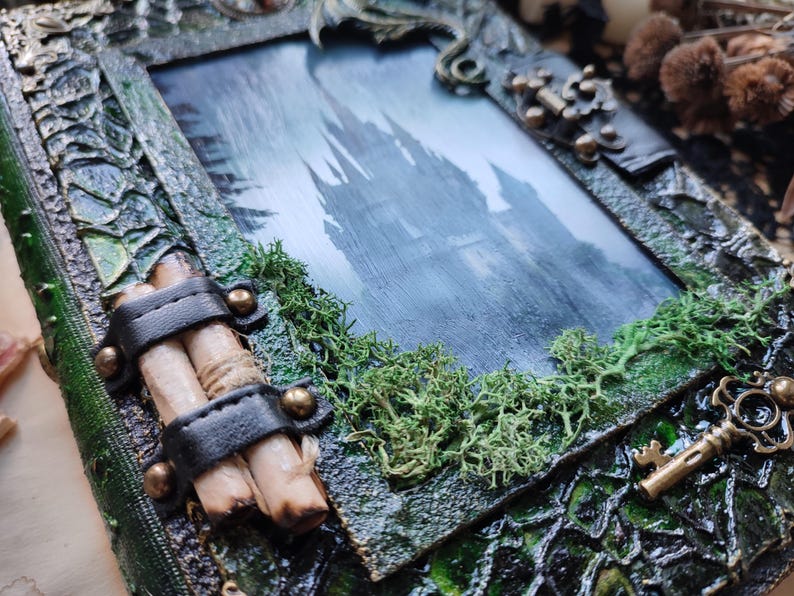 May include: A handcrafted, fantasy-themed picture frame with a castle scene. The frame is textured with green and brown tones, adorned with faux moss, a key charm, and a leather strap holding scrolls. The image inside depicts a castle.