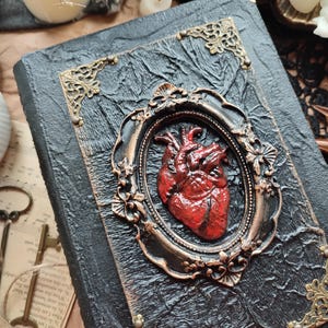 Pu&ograve; includere: Un diario rilegato in pelle nera con una cornice dorata ornata e un cuore anatomico rosso scolpito sulla copertina.
